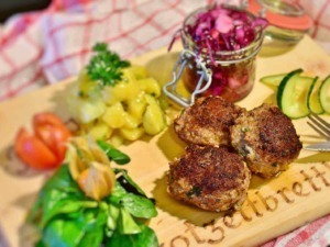 Fagottini di carne ripieni di formaggio e spezie, serviti con contorni di insalata, cetrioli, pomodorini e insalata di cavolo rosso, presentati su un tagliere di legno in stile rustico.