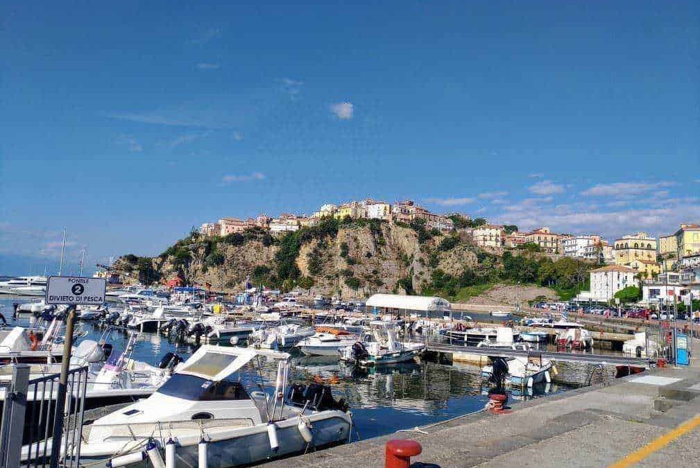 Pe scenari di un porto turistico con barche a vela e yacht, un paesino colorato su una scogliera, cielo azzurro con alcune nuvole.