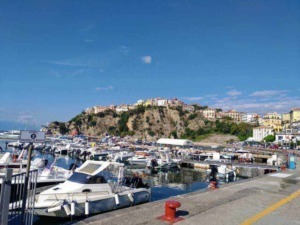 Pe scenari di un porto turistico con barche a vela e yacht, un paesino colorato su una scogliera, cielo azzurro con alcune nuvole.
