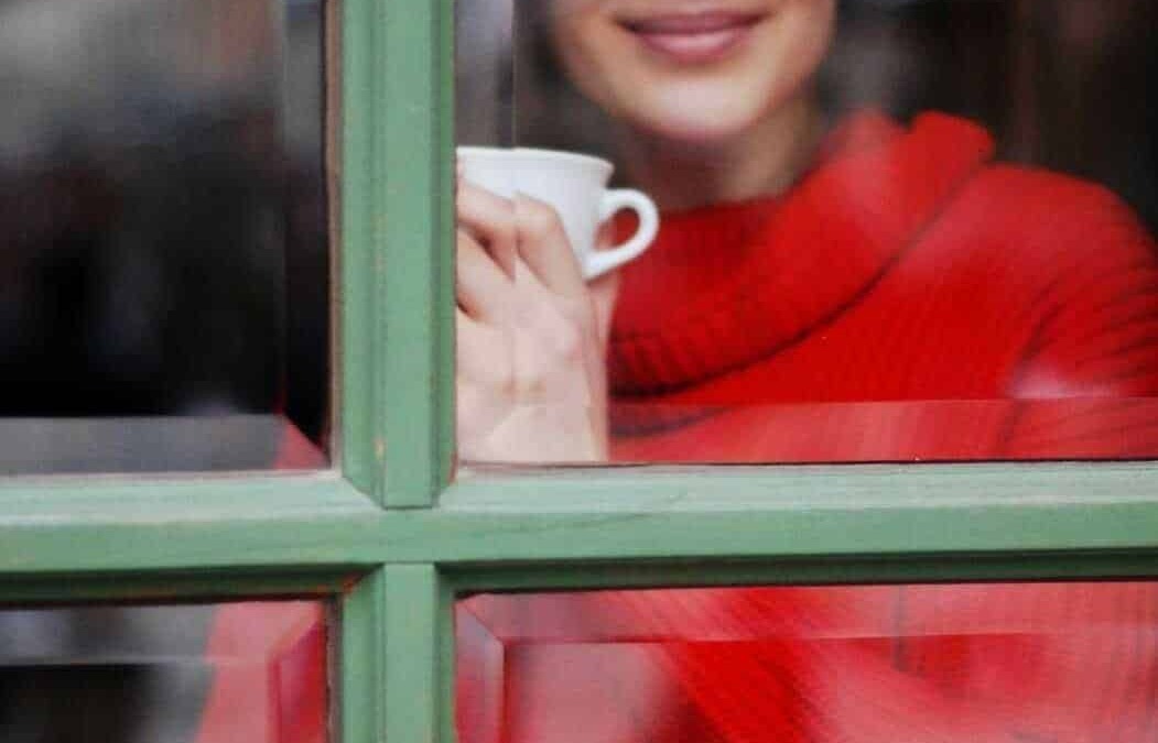 Tiziana sorridente guarda fuori da una finestra mentre sorseggia una tazza di caffè, creando un momento di relax e intimità in un ambiente accogliente.