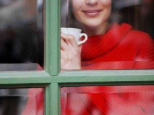 Tiziana sorridente guarda fuori da una finestra mentre sorseggia una tazza di caffè, creando un momento di relax e intimità in un ambiente accogliente.