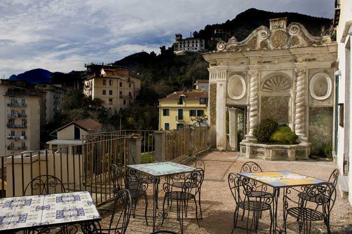 Balcone con vista sulla città e sulle montagne, tavoli all'aperto con sedie in ferro battuto, decorazioni architettoniche eleganti su un edificio storico in Italia.