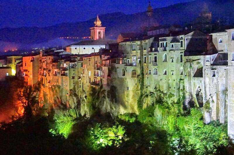 Palazzi colorati e case storiche su una scarpata, illuminati con luci verdi e arancioni, con sfondo di colline e cielo notturno, rappresentano un affascinante paesaggio urbano italiano.