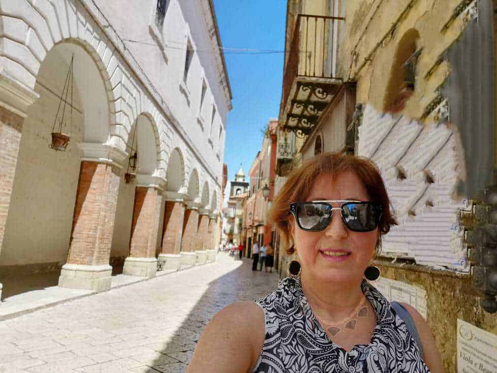 Scorcio di strada storica con arconi in mattoni e donna con occhiali da sole che prende un selfie a lume di sole.