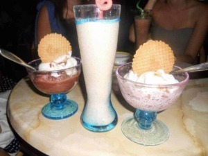 Vasche di gelato alla vaniglia con biscotti e un frappé alla fragola con decorazioni colorate, accompagnati da drink estivi in un ambiente festivo.