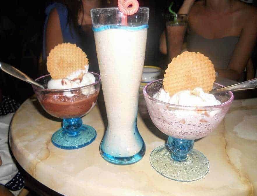 Vasche di gelato alla vaniglia con biscotti e un frappé alla fragola con decorazioni colorate, accompagnati da drink estivi in un ambiente festivo.