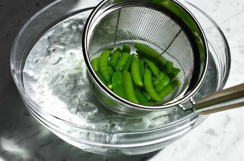 Verdure fresche di piselli verdi in una griglia metallica sopra ciotola con acqua ghiacciata per mantenere freschezza e colore.