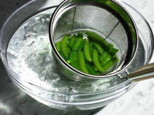 Verdure fresche di piselli verdi in una griglia metallica sopra ciotola con acqua ghiacciata per mantenere freschezza e colore.