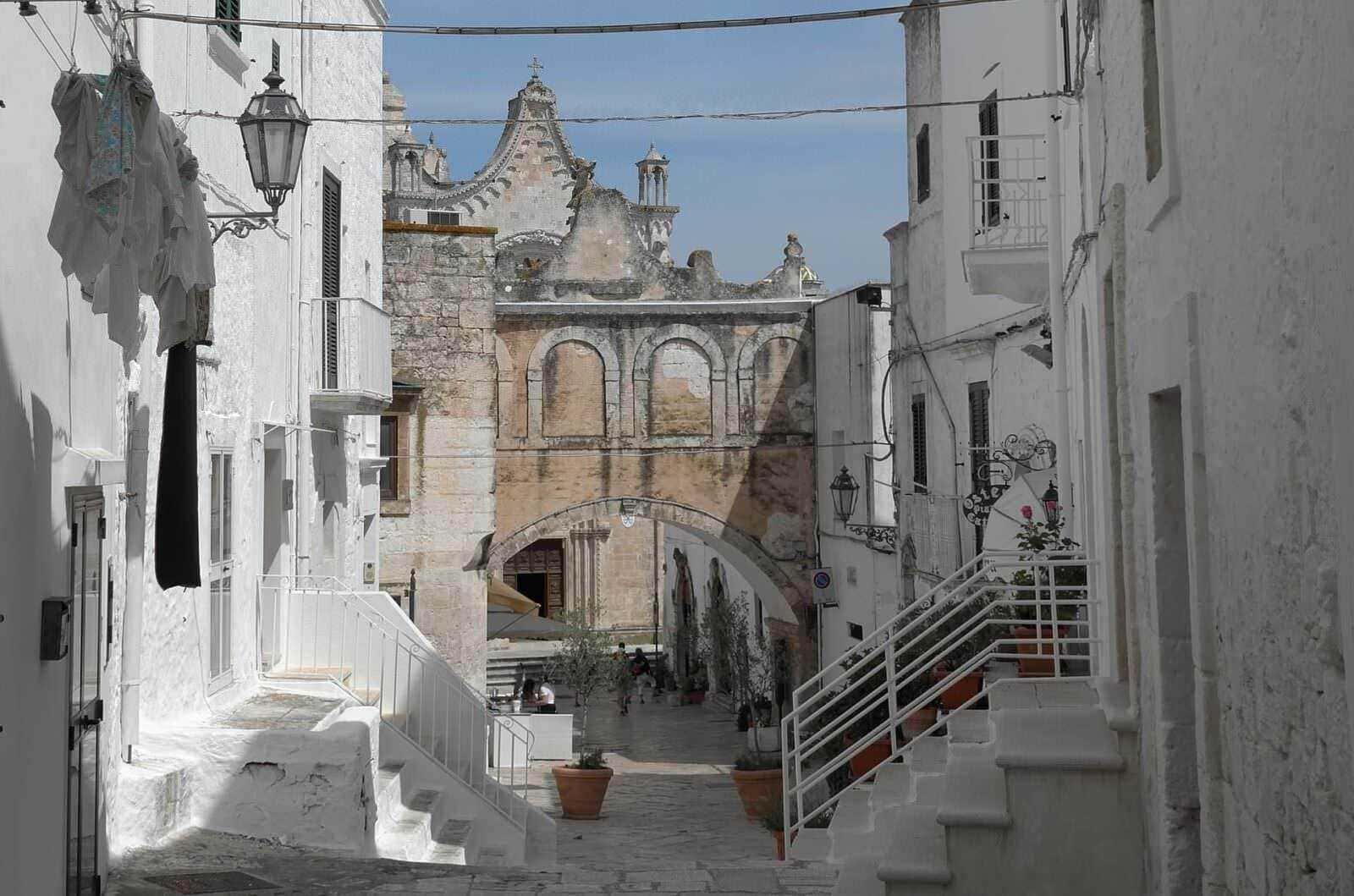 Viaggio in Puglia con Voyage Privé: 7 luoghi più belli da visitare 4 Vecchia strada di un paese in Puglia con mura bianche e archi, chiesa storica sullo sfondo, atmosfera mediterranea e architettura tradizionale.