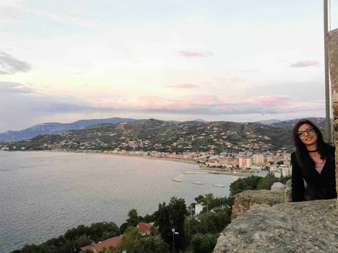 Panorama del mare e della costa al tramonto, con una donna sorridente seduta su un muro di pietra, sullo sfondo le colline e il cielo colorato