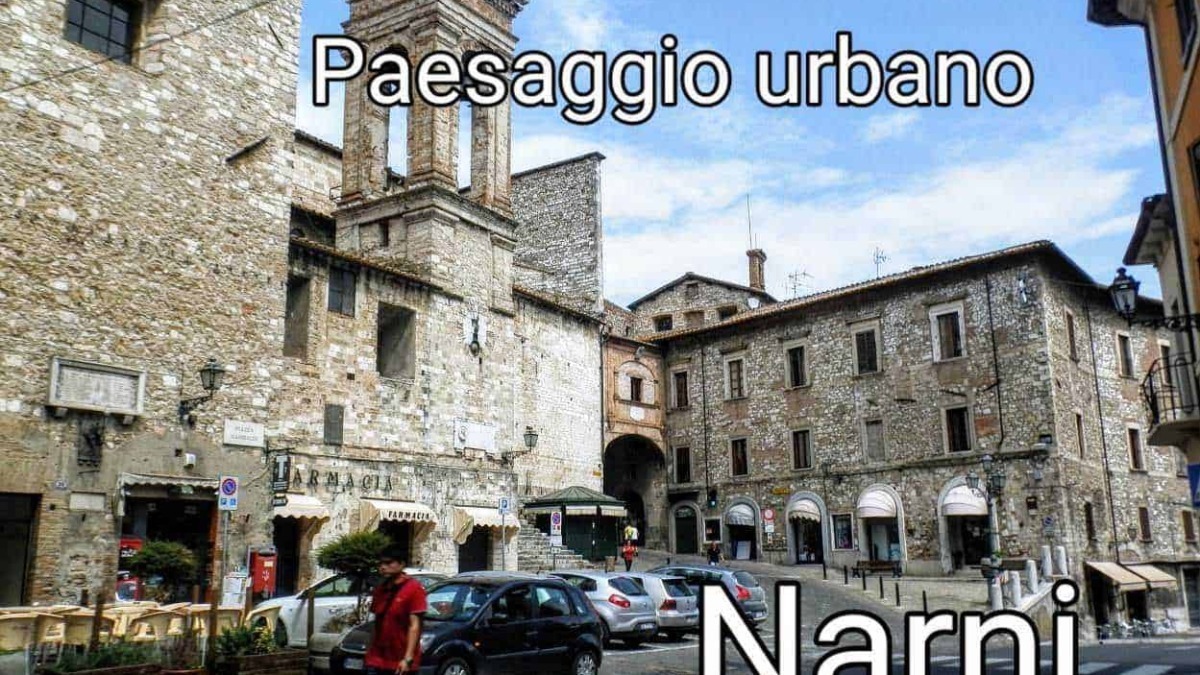 Vecchio centro storico di Narni, piazza con edifici in pietra, attività commerciali e uno skyline di antiche torri e mura medievali, rappresentativo di un paesaggio urbano ricco di storia e cultura.