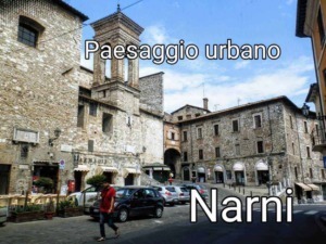Vecchio centro storico di Narni, piazza con edifici in pietra, attività commerciali e uno skyline di antiche torri e mura medievali, rappresentativo di un paesaggio urbano ricco di storia e cultura.