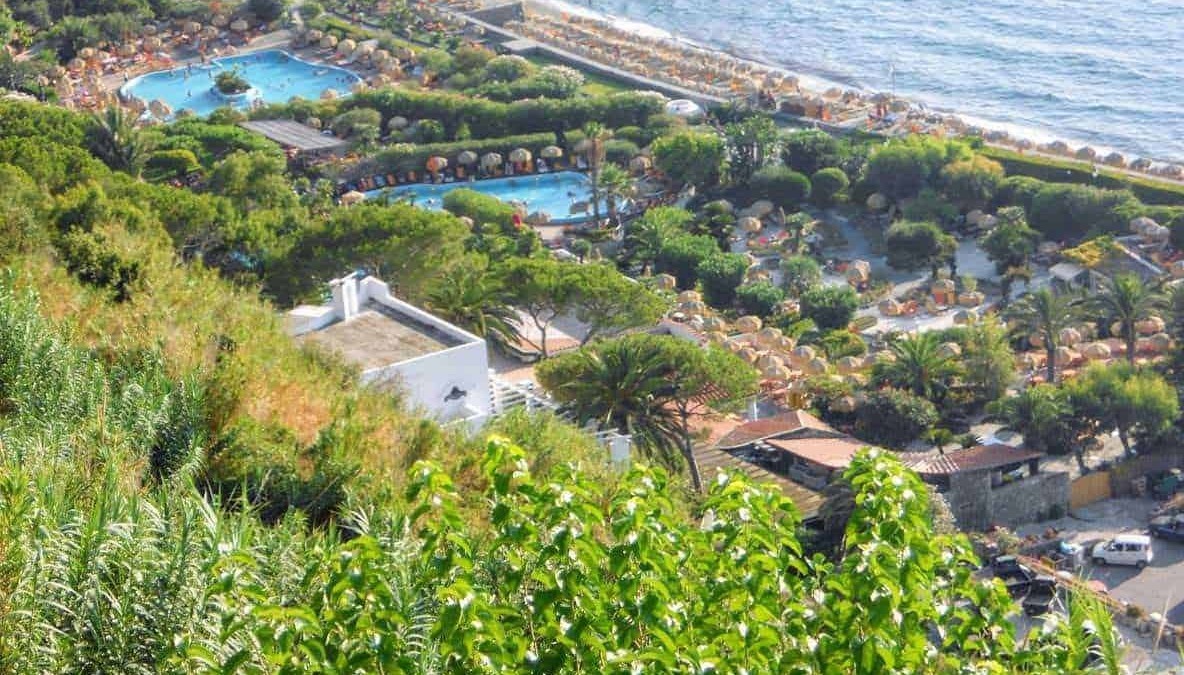 Spiaggia affacciata sul mare con piscine e vegetazione rigogliosa, vista dall'alto di una località turistica al mare in Italia.