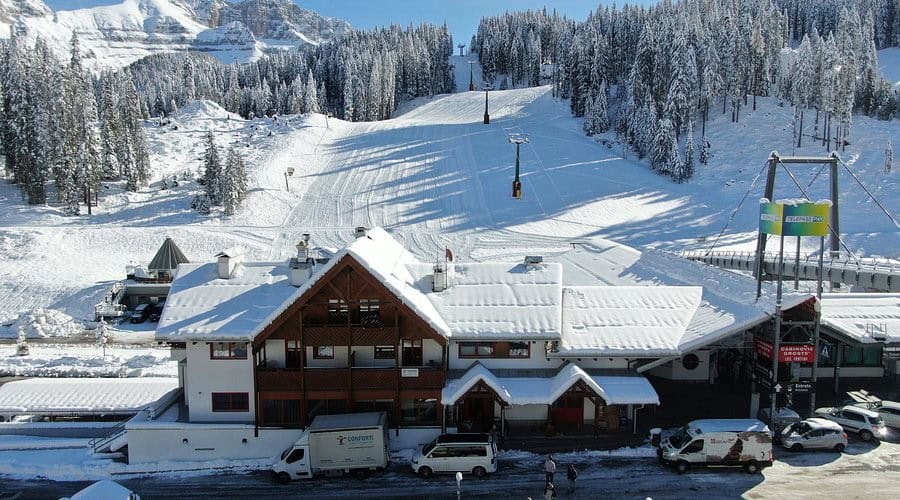Dormire in un Hotel a Madonna di Campiglio tra neve perenne e laghi azzurri 9 Immagine di un hotel a Madonna di Campiglio circondato da neve e boschi, con impianti di risalita e veicoli parcheggiati, ideale per chi cerca relax tra neve perenne e laghi azzurri.