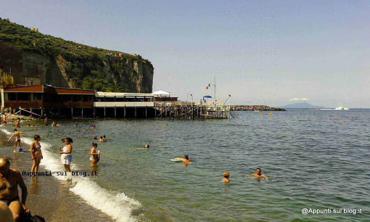 Spiaggia affollata con acqua cristallina, stabilimenti balneari e scogliera sullo sfondo, perfetta per relax estivi e attività acquatiche nel mare italiano.