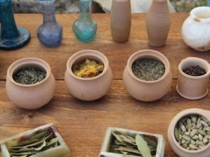 Fiori di erbe aromatiche e semi in terracotta su tavolo di legno, elementi naturali per la cucina e la salute, con bottiglie di vetro colorato sullo sfondo, rappresentando infusi e spezie fresche.