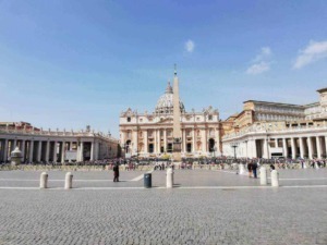 Città del Vaticano, Piazza San Pietro con la basilica e obelisco al centro, affollata di visitatori e turisti, cielo sereno, esempio di arte e architettura religiosa.