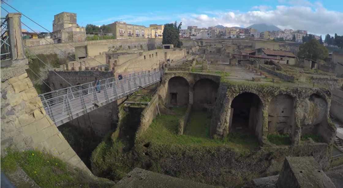 Antica area archeologica con rovine romane e struttura moderna di passerella in metallo, immersa in un contesto urbano, perfetta per appunti sulla storia e l'archeologia.