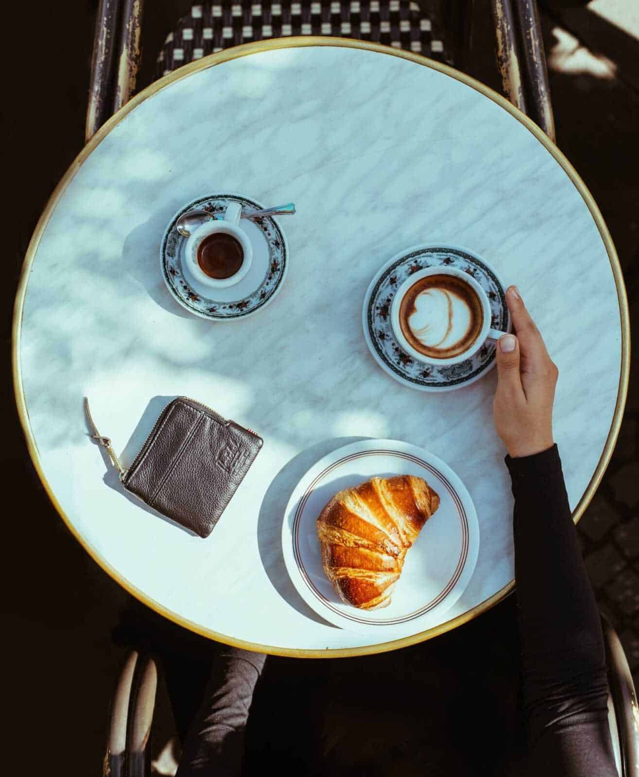 Una tazza di cappuccino con latte decorato, una tazzina di caffè, un croissant dorato e una borsetta su un tavolo in marmo di un bar elegante, perfetto per appunti sul blog.