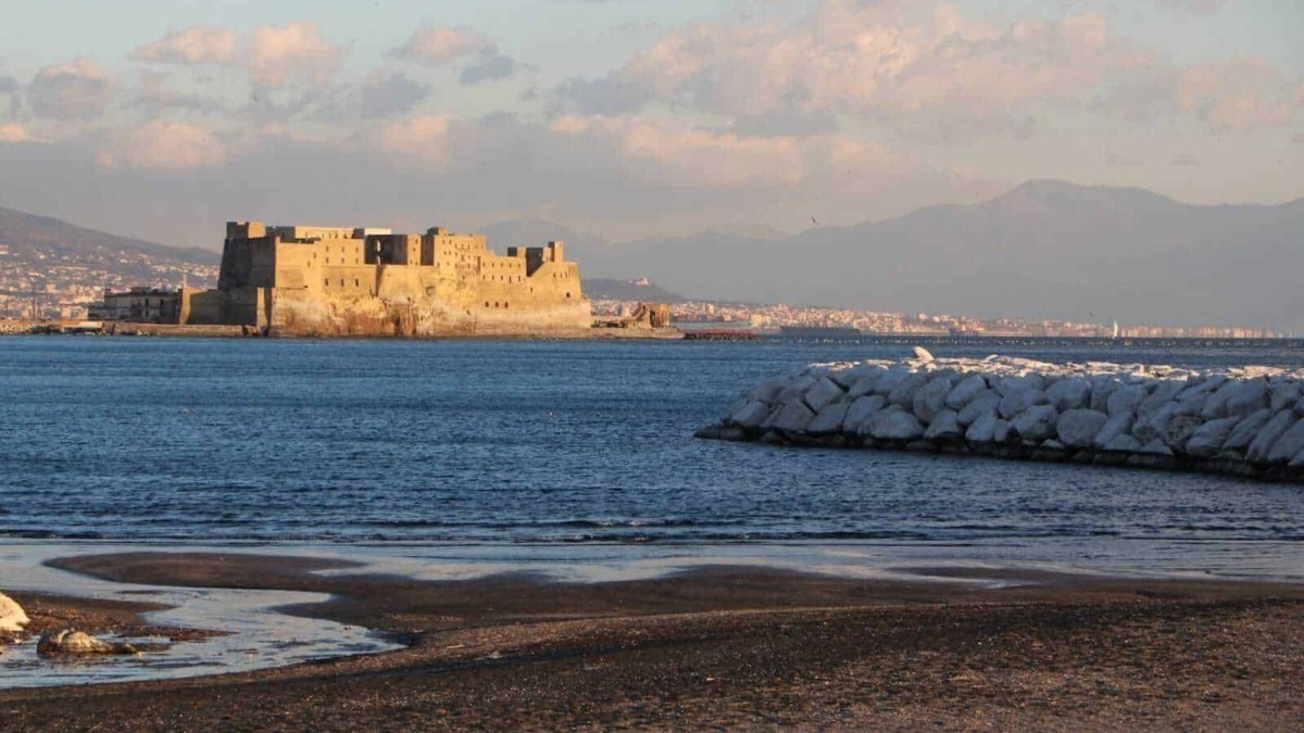 Castello Aragonese di Ischia sul mare al tramonto, vista panoramica delle influenze storiche e culturali dell’isola di Ischia, in Campania, Italia, con sfondo di montagna e cielo nuvoloso.