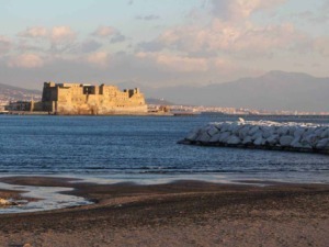 Castello Aragonese di Ischia sul mare al tramonto, vista panoramica delle influenze storiche e culturali dell’isola di Ischia, in Campania, Italia, con sfondo di montagna e cielo nuvoloso.