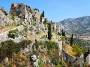 Antico castello medievale sulla collina, con mura in pietra e torri, circondato da cactus e cipressi, panoramica sulle montagne e il paesaggio sottostante.