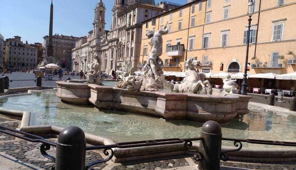 Fontana storica con sculture di marmo nel centro di Roma, con la chiesa e Palazzo sullo sfondo, in una giornata soleggiata, zona turistica e di ritrovo.