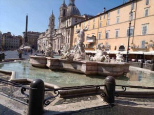 Fontana storica con sculture di marmo nel centro di Roma, con la chiesa e Palazzo sullo sfondo, in una giornata soleggiata, zona turistica e di ritrovo.