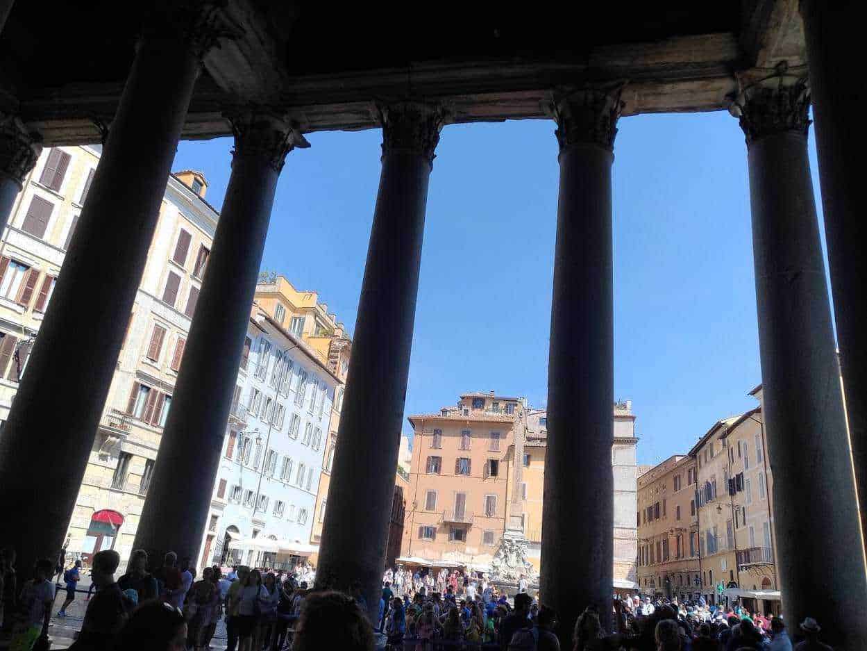 Immagine di un antico foro romano con colonne di granito, illuminato dal sole, affollato di visitatori, con palazzi storici italiani sullo sfondo, rappresenta il cuore della città di Roma.