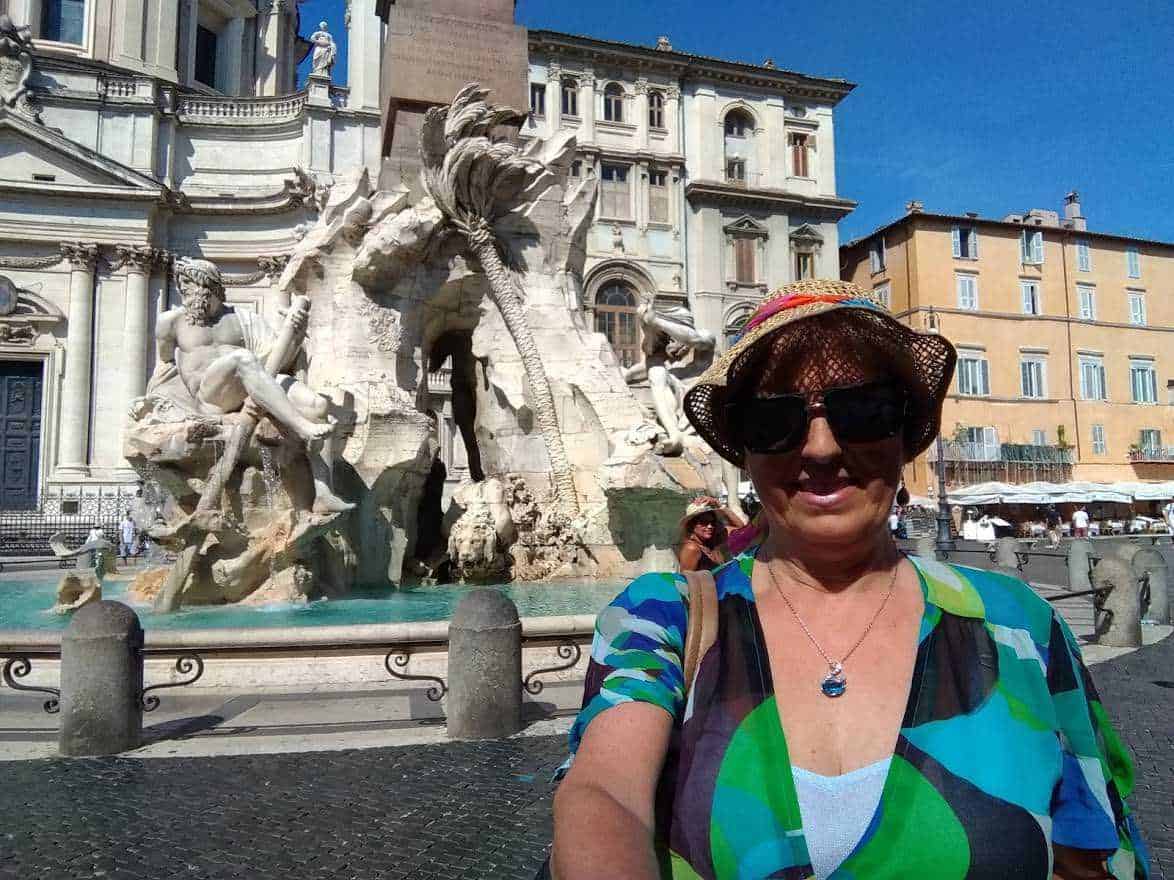 Fonte: Fontana dei Quattro Fiumi a Piazza Navona, Roma, con donna sorridente con cappello e occhiali scuri in primo piano.
