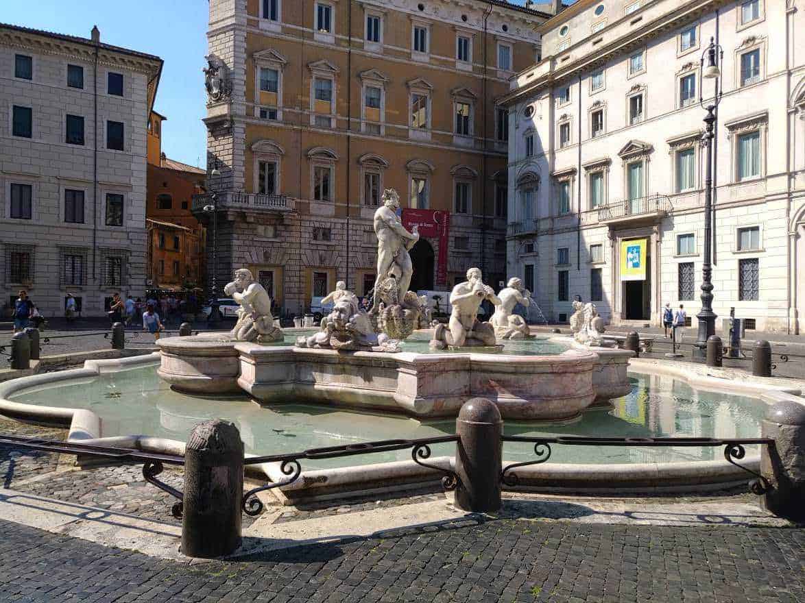 Fontana storica con sculture in pietra situata in una piazza di Roma, circondata da edifici storici e architettura classica, ideale per foto turistiche e passeggiate urbane.