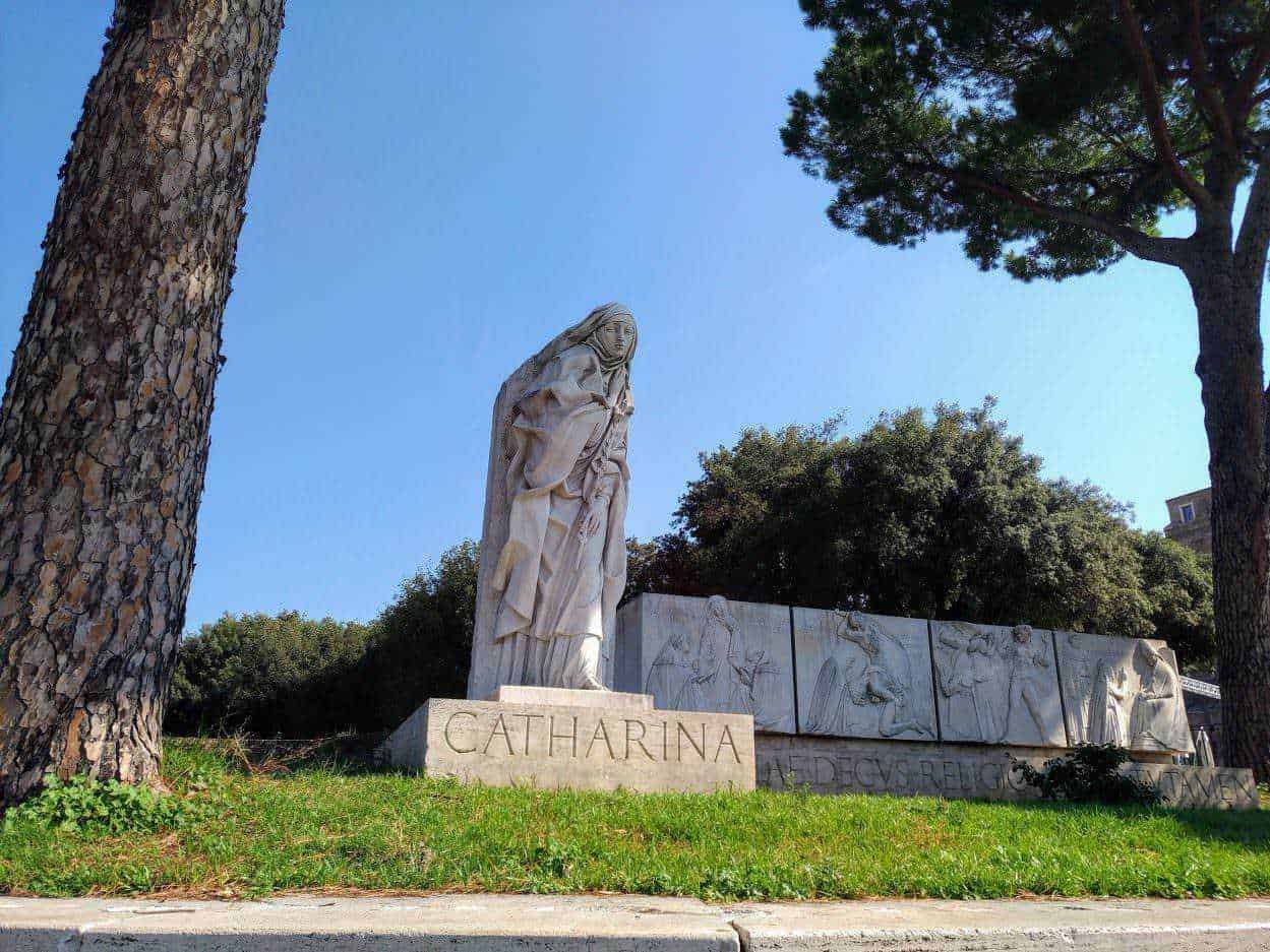 Statua di Santa Caterina in marmo con sfondo di alberi e cielo azzurro, monumento storico situato in un parco cittadino, omaggio alla santa con dettagli scolpiti e iscrizione sulla base.