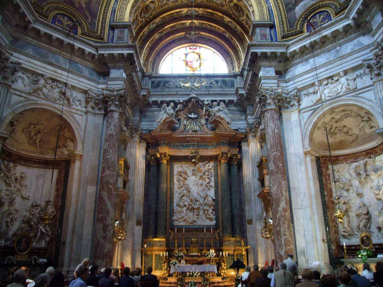 Sant'Agnese in Agone a Roma: luoghi da vedere in stile barocco 3 Maestoso interno barocco con decorazioni dorate, colonne in marmo e soffitto affrescato, chiesa storica con altare centrale, affluenza di fedeli, visitatori e dettagli artistici.