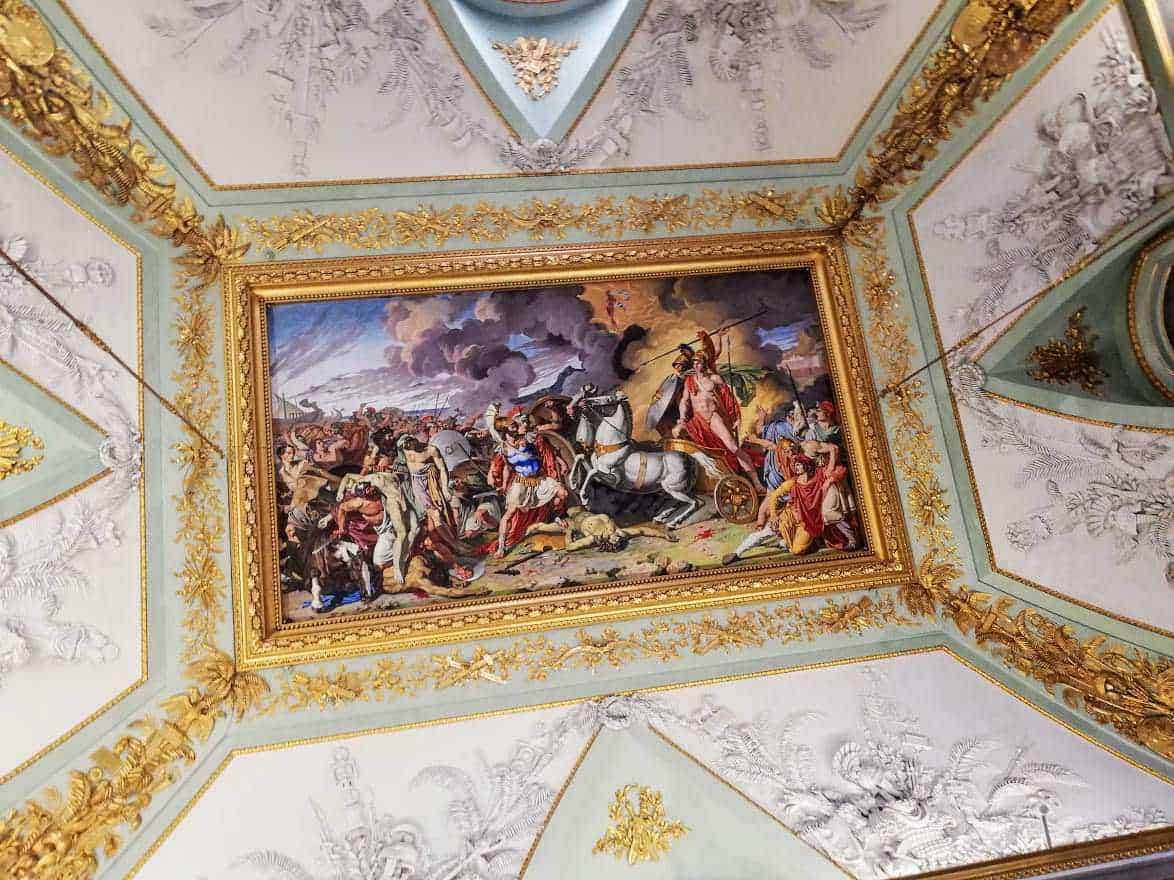 Dipinto storico che rappresenta una battaglia epica, incorniciato da un soffitto decorato con dettagli dorati e ornamentali, situato in un palazzo o museo artistico di prestigio.