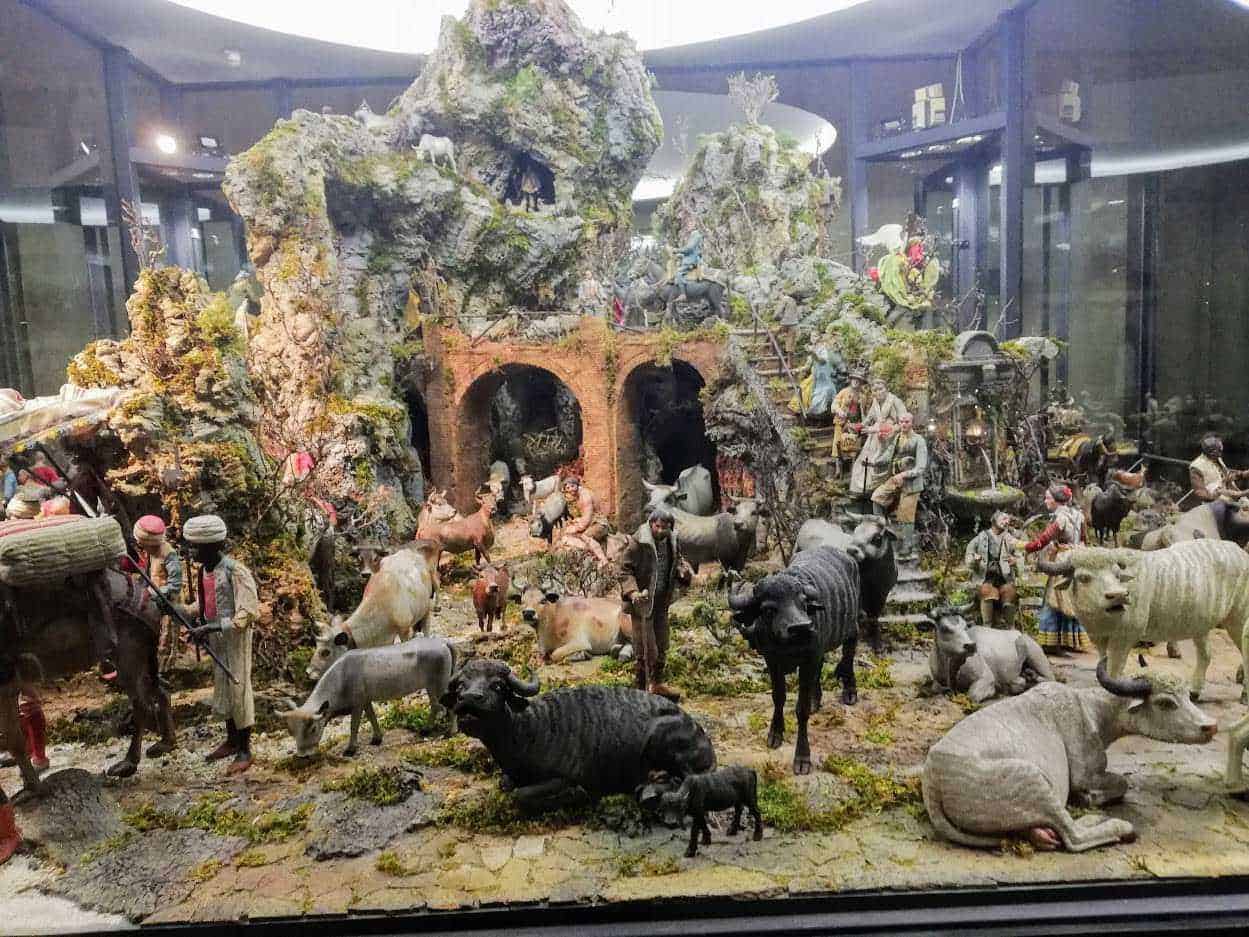 Rilievo di presepe con figure di animali e persone in stile tradizionale, ambientato in un paesaggio montano con archi in mattoni, piante verdi e figurine artigianali, rappresentazione natalizia dettagliata e ricca di atmosfera.