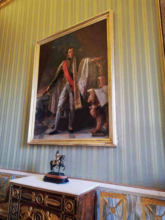 Ritratto storico di un ufficiale in stile neoclassico, decorato con dettagli eleganti e pose regali. L'opera si trova in una cornice dorata su una parete con carta da parati a righe verdi e oro. Sotto, mobile antico con statuetta e decorazioni sofisticate.