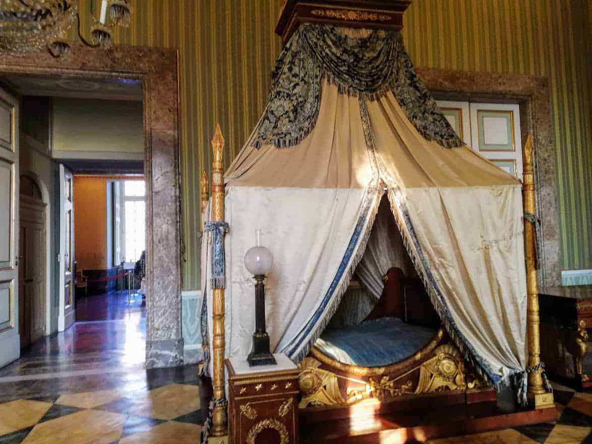 Antico letto a baldacchino in stile barocco in una stanza storica, decorato con drappeggi e dettagli dorati, all'interno di un palazzo d'epoca con pareti rivestite in velluto e pavimento in parquet.