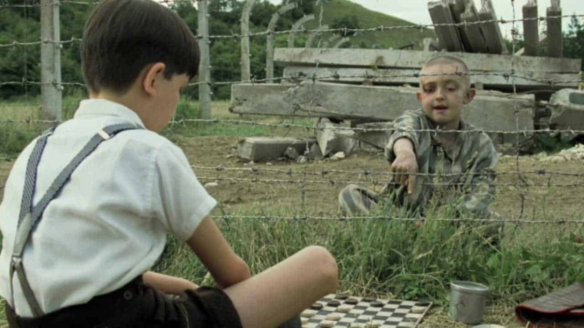 Un bambino e un ragazzo giocano a scacchi all'aperto dietro una recinzione di filo spinato in un contesto di rovina e desolazione.