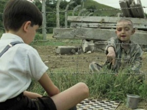 Un bambino e un ragazzo giocano a scacchi all'aperto dietro una recinzione di filo spinato in un contesto di rovina e desolazione.
