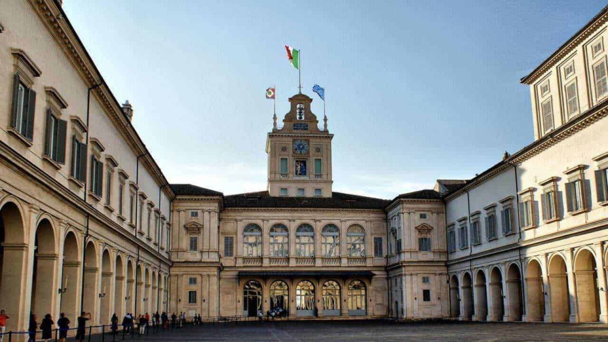 Storico palazzo con architettura classica, facciata elegante e torre con bandiere italiane e europee, in un cortile di pietra con persone in fila, ambientazione urbana e monumentale.