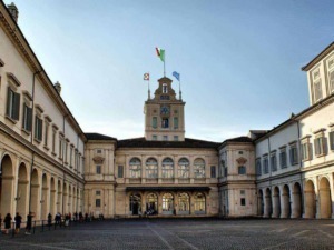 Storico palazzo con architettura classica, facciata elegante e torre con bandiere italiane e europee, in un cortile di pietra con persone in fila, ambientazione urbana e monumentale.