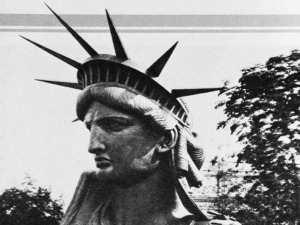 Statua della Libertà in stile vintage, foto in bianco e nero, simbolo di libertà e democrazia, raffigurante una donna con diadema a raggi. Scena urbana con alberi sullo sfondo, atmosfera retrò.
