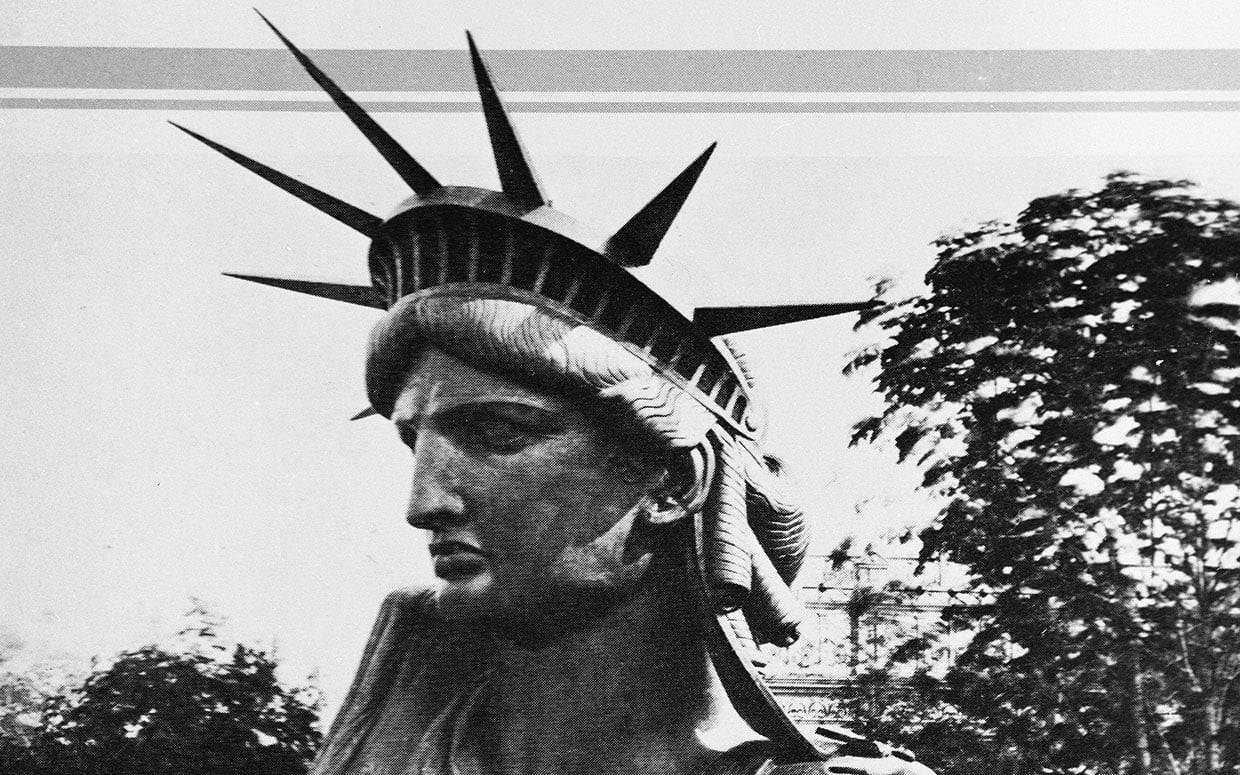 Statua della Libertà in stile vintage, foto in bianco e nero, simbolo di libertà e democrazia, raffigurante una donna con diadema a raggi. Scena urbana con alberi sullo sfondo, atmosfera retrò.