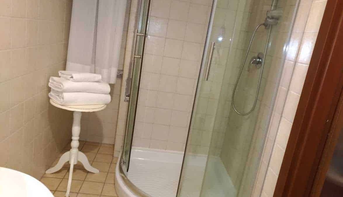 Accappatoi da bagno bianchi ordinatamente piegati su un tavolino in un bagno con doccia semicircolare, piastrelle beige e armadietto in legno, ambientazione moderna e pulita.