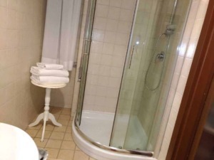 Accappatoi da bagno bianchi ordinatamente piegati su un tavolino in un bagno con doccia semicircolare, piastrelle beige e armadietto in legno, ambientazione moderna e pulita.