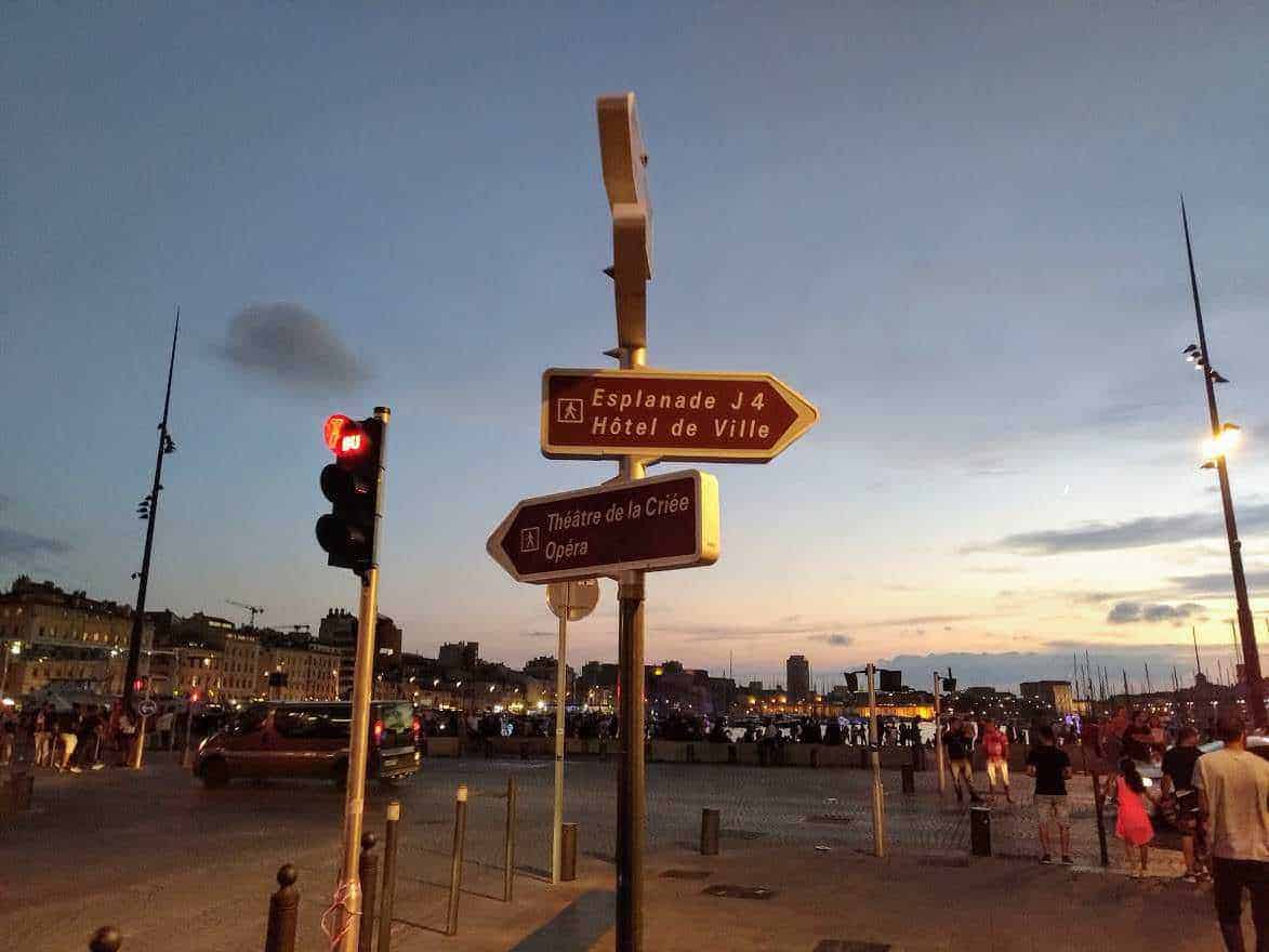Cosa vedere a Marsiglia a piedi dal porto di sera 5 Palo con segnali stradali di indicazione a Marsiglia al tramonto, con il porto e le persone che passeggiano sul lungomare. Scena urbana vivace di sera con cielo al crepuscolo, ideale per blog di viaggio e turismo.