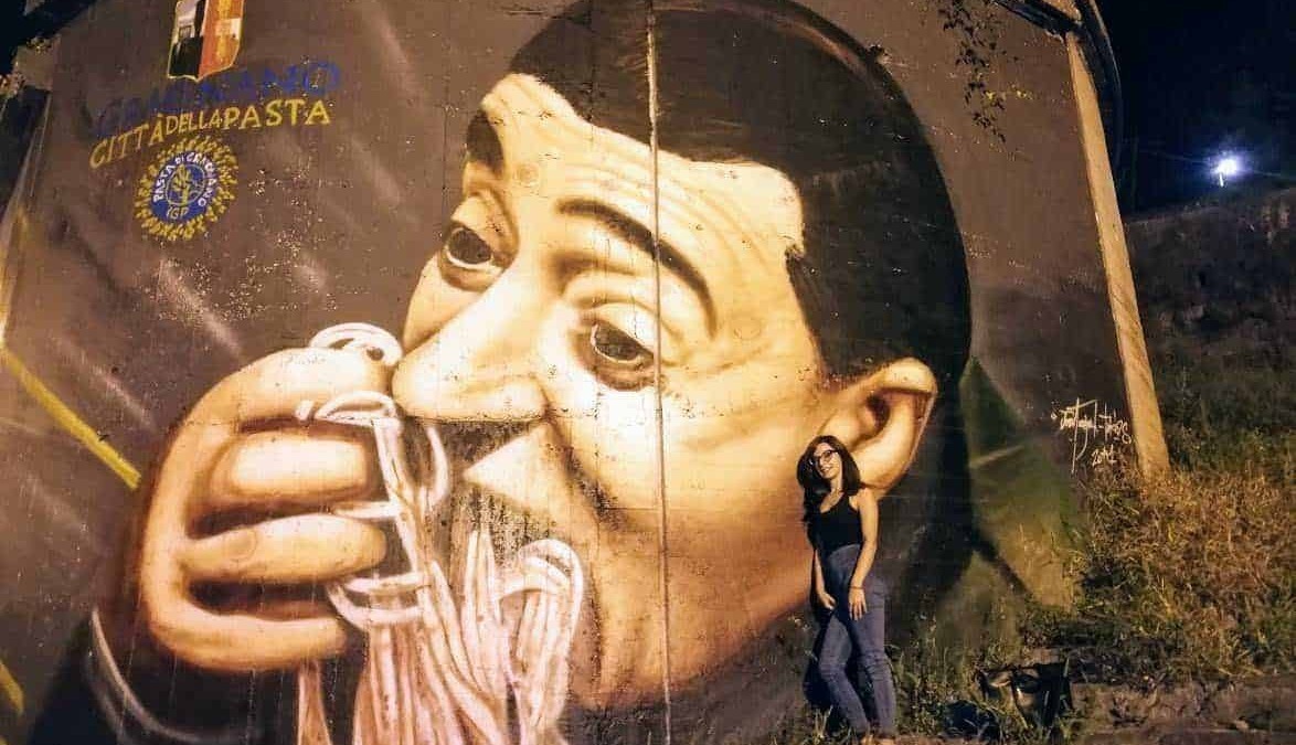 Vignetta di graffiti artistico rappresentante un uomo che sorseggia uno spaghetti, situato sul muro di una città italiana, con una donna in piedi accanto, sotto un cielo notturno.