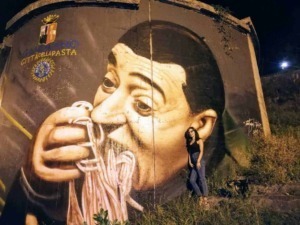 Vignetta di graffiti artistico rappresentante un uomo che sorseggia uno spaghetti, situato sul muro di una città italiana, con una donna in piedi accanto, sotto un cielo notturno.