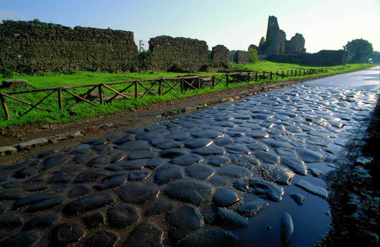 Antico camino di pietra con rovine romane sullo sfondo, sotto un cielo blu, immerso in un paesaggio verde naturale, ideale per esplorare storia e natura.
