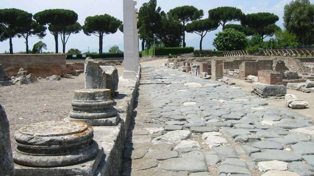 Antico sito archeologico di Roma antica con pavimentazione in pietra e resti di colonne, immerso nel verde di pino marittimo e cipressi, esempio di archeologia e storia romana.